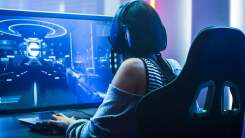 A woman sits at a computer playing a game while wearing a headset