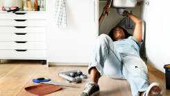 Photo of a Black woman wearing light wash jeans and a light blue shirt lying down on her back to fix the kitchen sink.