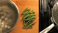 A bowl of ice water next to trimmed green beans and a pot of boiling water.
