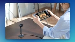 Nintendo Switch 2 Camera accessory on a brown desk, connected to a Switch 2, held by a woman who's playing a game on the console.