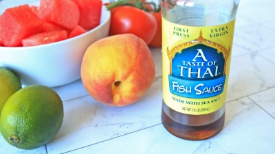 A bottle of fish sauce next to a bowl of cut watermelon, limes, and other fruit.