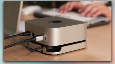 Satechi's M4 Mac mini dock, sitting against the base of the Mac mini on a brown wooden desk with a person's hands visible in the background, typing on a keyboard.