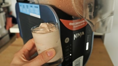 A hand holding a milkshake in front of the Ninja Slushi machine