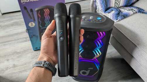 My hand holding the Soundcore Rave 3S microphones in my office with the Soundcore Rave 3S speaker in the back.