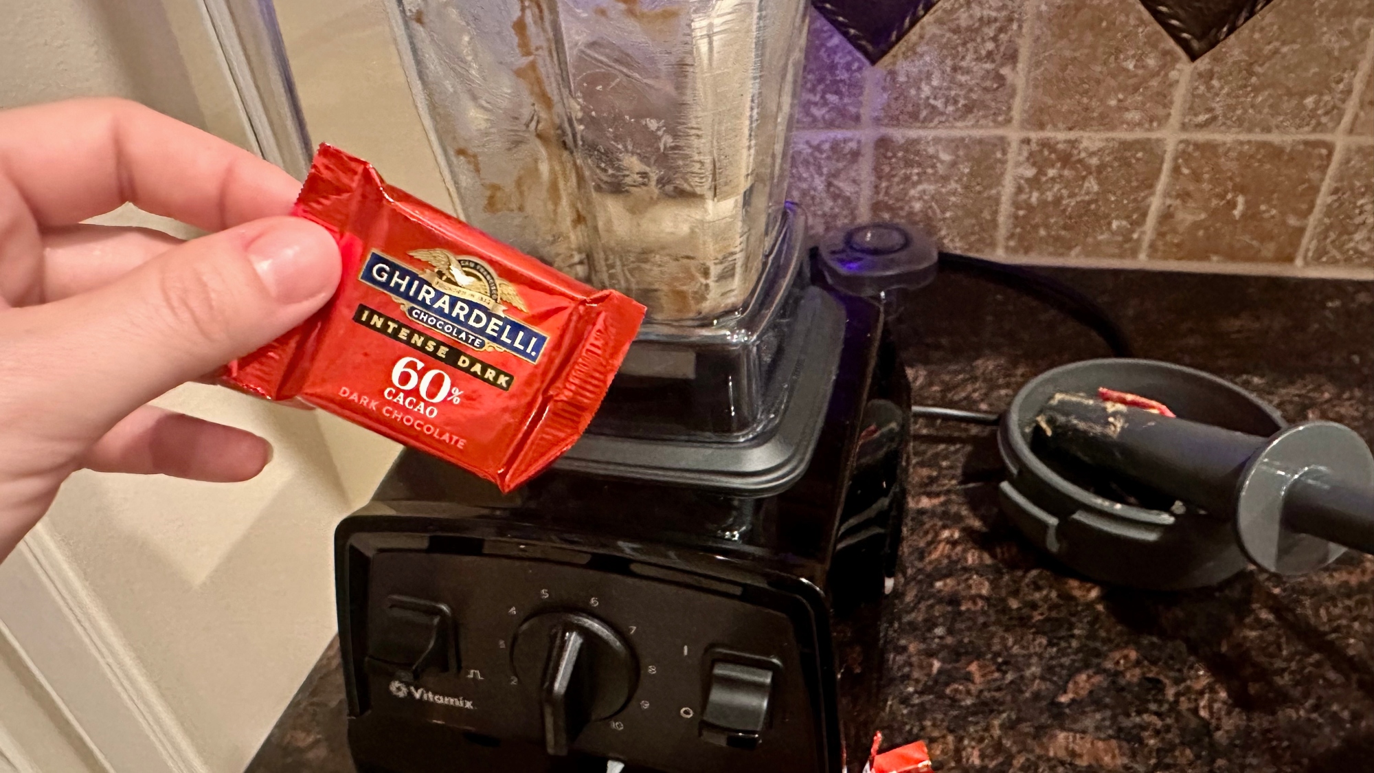 A hand holding a chocolate square in front of blender.