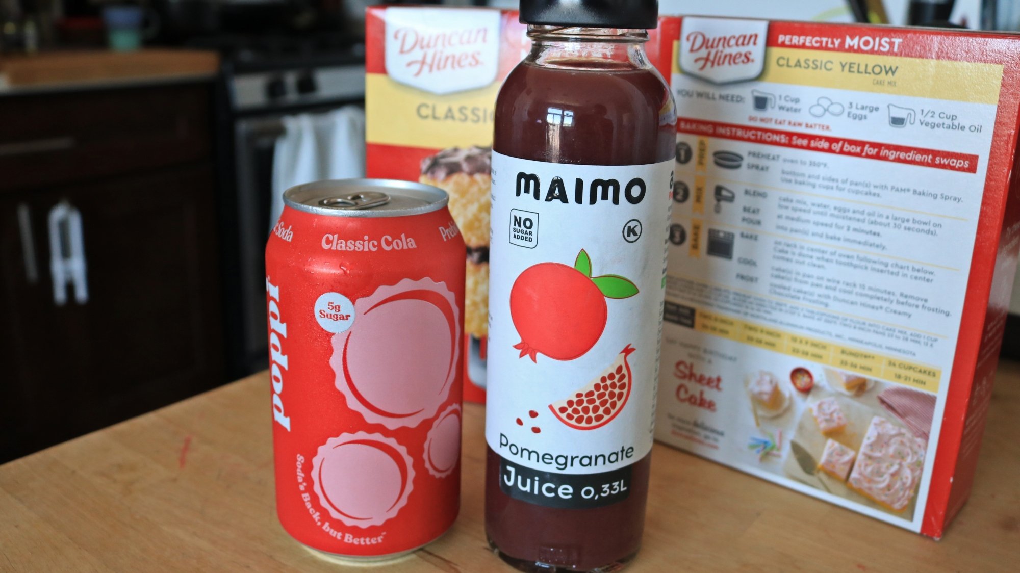 A can of cola and a bottle of juice in front of two boxes of cake mix.