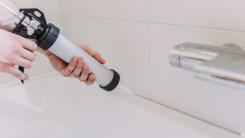 hands holding a caulking gun and applying silicon caulk to a tub in a bathroom
