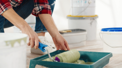 Person applying paint to a roller in a tray, surrounded by other painting accessories and tools