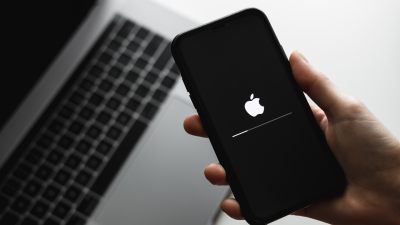 A hand holding an iPhone running a software update, with an open MacBook visible in the background