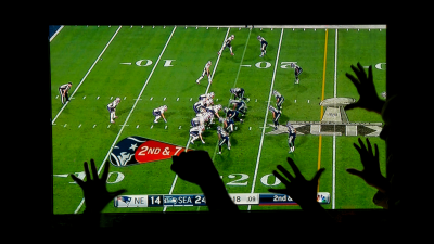 The Super Bowl playing on a flatscreen TV in a dark room with viewers visible cheering in front of it