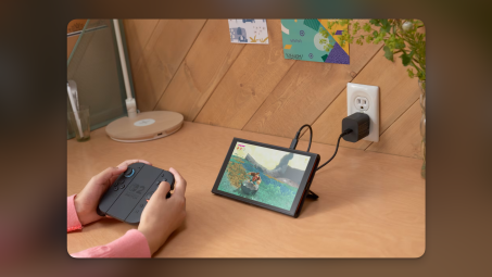 A Nintendo Switch 2 connected to a wall charger, with a person playing a game using the Joy-Con controllers.